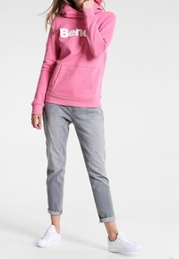 Sudadera rosa con grandes letras blancas que dicen "Bend," presenta un bolsillo tipo canguro. Combinada con jeans claros grises remangados y zapatillas blancas.