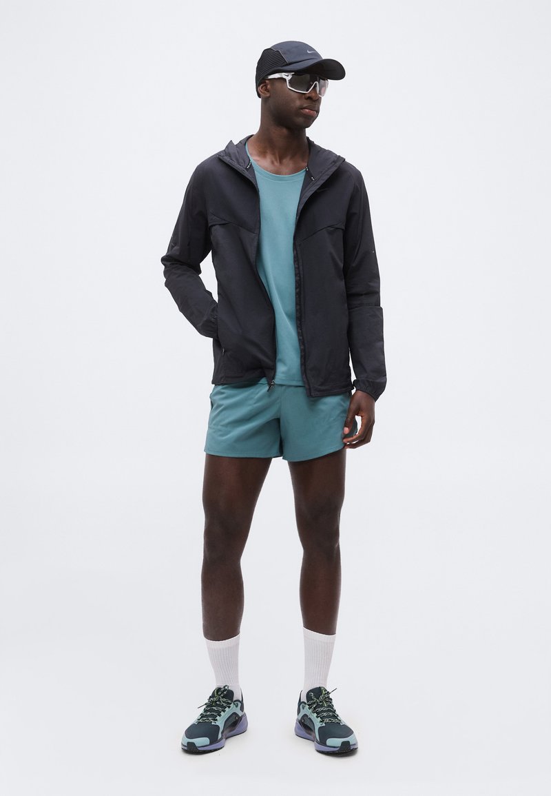 Hombre lleva una camiseta y pantalones cortos deportivos color verde azulado, chaqueta negra con cremallera, gorra negra, calcetines blancos, zapatillas de correr verde azulado y negras, y gafas de sol deportivas blancas.