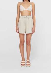 Vrouw in een witte kanten bralette, beige high-waist short met trekkoord, en crèmekleurige sandalen, staande tegen een effen lichte achtergrond.