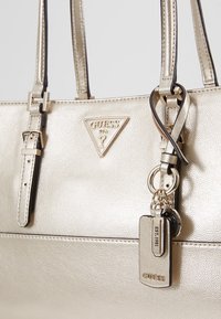 Kultainen metallinhohtoinen käsilaukku, jossa on teksturoidu pintaa, kaksi hihnaa, kolmion muotoinen logo ja vetoketjuun kiinnitetyt avaimenperäkoristeet.