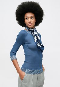 Afro frizurás nő, kék csipkés topban, csíkos nyakkendőben és világosszürke csíkos nadrágban, kezét a zsebében tartja.