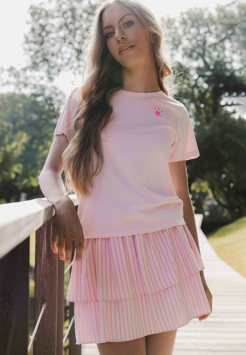 Lichtroze t-shirt met korte mouwen, voorzien van een klein roze logo. Daaronder een geplooide rok met afwisselend effen en gestreepte lagen.