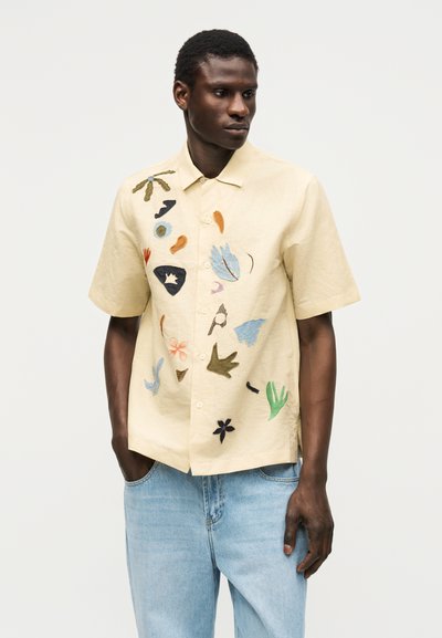 Άνδρας φοράει μπεζ κοντομάνικο πουκάμισο με πολύχρωμα αφηρημένα κεντητά σχέδια, σε συνδυασμό με ανοιχτό μπλε τζιν, στέκεται με τα χέρια στις τσέπες.