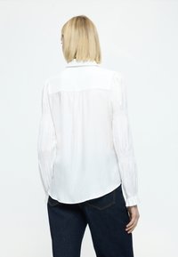 Bílá halenka s dlouhými nařasenými rukávy, zadním dílem a hladkou, plynulou texturou. Ve spojení s tmavě modrými džínami.