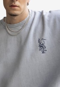 Felpa grigia chiaro realizzata in misto cotone, con una grafica di un airone blu navy e ricami di foglie sul lato sinistro del petto. Visibile una collana a doppia catena.