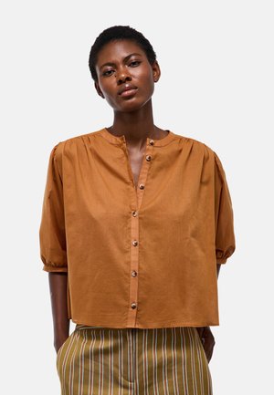 Femme portant un chemisier marron ample à boutons avec des manches bouffantes et un pantalon rayé, debout les mains dans les poches devant un fond uni.