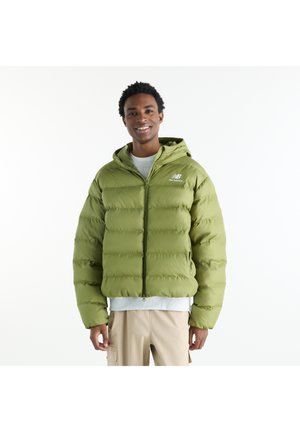 Mann lächelt und trägt eine grüne New Balance Steppjacke mit Kapuze, hellgraues Shirt und beige Hose vor weißem Hintergrund.