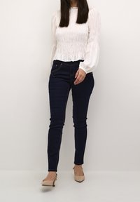 Bílý smockový top s dlouhými nabíranými rukávy, zkombinovaný s tmavě modrými skinny džíny a béžovými lodičkami s špičatou špičkou, stojící na neutrálním pozadí.