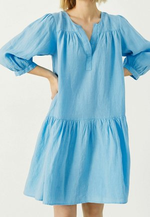 Femme portant une robe ample bleu clair à manches longues avec des détails froncés au niveau de l'encolure et de l'ourlet, debout les mains sur les hanches.