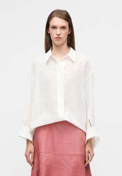 Blouse blanche transparente à boutons avec un motif à pois texturé, col pointu et manches larges. Assortie à une jupe en cuir rose.