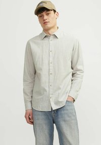 Camicia a maniche lunghe, grigia chiaro a righe, con frontale a bottoni e colletto, abbinata a jeans azzurri e un cappellino beige con un logo.