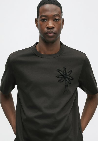 Tricou negru din bumbac cu un design brodat de floare pe partea stângă a pieptului. Mâneci scurte și decolteu rotund. Textură netedă.