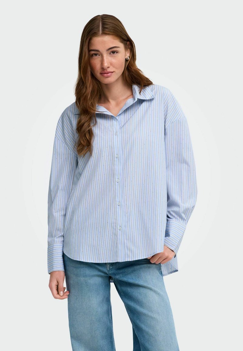 Jeune femme aux longs cheveux bruns portant une chemise à boutons rayée bleu clair et un jean bleu sur un fond blanc uni.