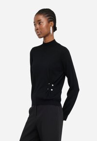 Maglione nero con collo alto, texture in maglia morbida e dettagli laterali con due perle. Abbinato a pantaloni scuri.