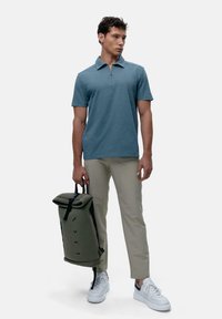 Téglalapú zöld póló rövid ujjakkal, világos bézs nadrágok és sötétzöld, felgöngyölhető hátizsák. Fehér sportcipők texturált részletekkel.