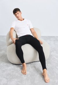 Černé legíny s přiléhavým střihem a páskem s logem, nošené s bílým tričkem. Modelka sedí na texturované krémové židli.