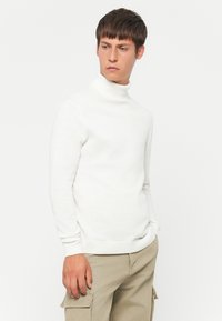 Joven vestido con un suéter blanco de cuello alto y pantalones cargo beige, de pie con una mano en el bolsillo, mirando de lado contra un fondo blanco liso.