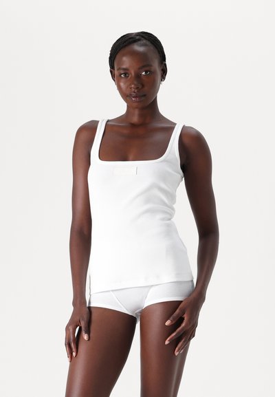 Débardeur blanc à côtes avec un décolleté carré et une petite étiquette sur le devant, associé à un shorty blanc à côtes. Tissu doux et coupe ajustée.