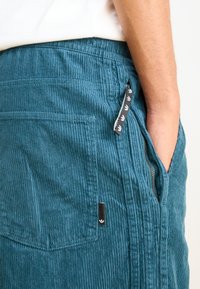 Teal corduroy pants with a textured surface, featuring a side zip pocket and a small black logo patch. The design includes horizontal ribbing.