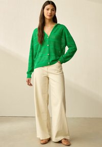 Grön stickad cardigan med V-hals, texturerat mönster och knappstängning. Kombinerad med krämfärgade vida byxor och neutrala skor.