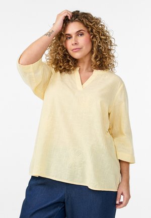 Femme aux cheveux bouclés portant un chemisier ample jaune pâle à col en V avec des manches trois-quarts et un pantalon bleu foncé, posant avec la main près des cheveux.