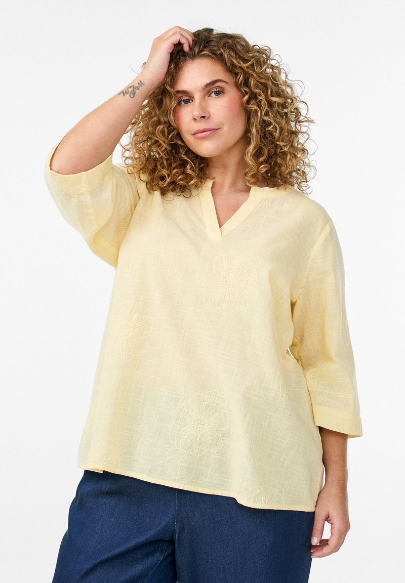 Mujer de cabello rizado con una blusa suelta amarilla pálida de cuello en V y mangas tres cuartos, y pantalones azul oscuro, posando con la mano cerca del cabello.