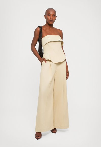 Top beige sem alças com uma bainha peplum curvada, combinado com calças de perna larga. Os detalhes incluem um botão no top e uma alça escura texturizada.