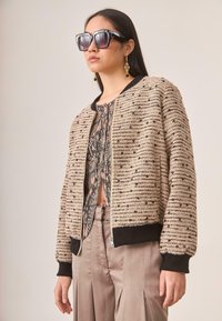 Cazadora bomber beige texturizada con ribetes negros, que cuenta con un cierre delantero y una blusa interior con estampado. Combinada con pantalones de satén marrón claro.