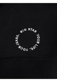 Text alb brodat, care spune „BIG STAR YOUR LIFE. YOUR JEANS.”, aranjat într-un cerc pe țesătură neagră.