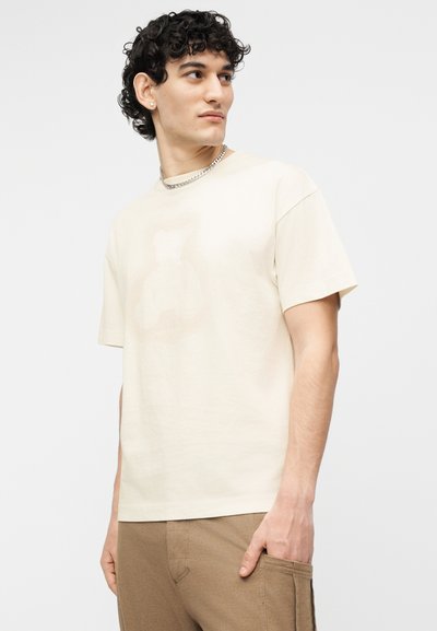 Νέος άνδρας με σγουρά μαλλιά, που φοράει μια απλή κρεμ κοντομάνικη μπλούζα, μπεζ παντελόνια, ασημένιο αλυσίδα λαιμού και σκουλαρίκι, κοιτάζοντας προς το πλάι.