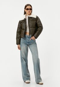 Bruine nep leren cropped jack met witte nep bontkraag, gecombineerd met lichtblauwe wijdetopje jeans en witte sneakers. Zwarte zonnebril.