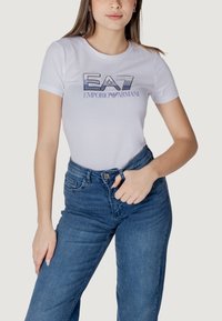 Maglietta bianca aderente con il logo "EA7 EMPORIO ARMANI" blu sul petto, abbinata a jeans blu a vita alta. Tessuto con texture liscia.