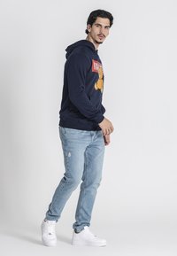 Sudadera marina con un gráfico de un oso, combinada con jeans claros desgastados y zapatillas blancas, de pie contra un fondo blanco.