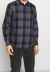 Chemise à manches longues à carreaux dans des tons bleu foncé, noir et jaune. Elle présente un col boutonné et une poche poitrine avec un détail de marque.