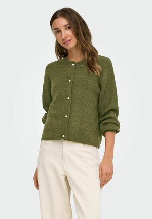 Femme portant un cardigan vert boutonné avec des boutons en forme de perles et un pantalon blanc cassé, debout, une main détendue le long du corps.