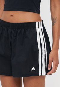 Melni sporta šorti ar elastīgu jostu, kuriem katrā pusē ir trīs baltas vertikālas svītras un balts Adidas logos apakšmalā.