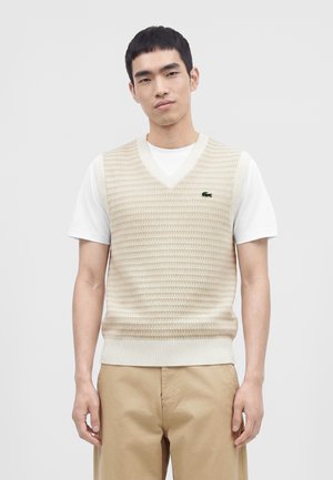 Jeune homme portant un gilet en maille beige par-dessus un t-shirt blanc et un pantalon beige, debout devant un fond clair uni.
