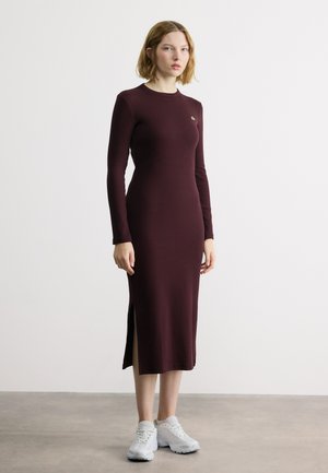 Femme debout portant une robe midi à manches longues de couleur bordeaux foncé avec une fente latérale et des baskets blanches, sur un fond clair uni.