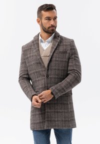 Ruskean ruutukuvioinen villa takki, joka on varustettu huivikauluksella, yksittäisellä nappisuljennalla ja etutaskuilla. Se on kerrostettu beige neuleen ja valkoisen paidan päälle.