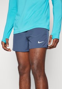 Tummansininen Nike-shortsit, joissa on valkoinen logo sivulla, yhdistettynä pitkähihaiseen turkoosiin puseroon, joka esittelee kevyen kankaan ja urheilullisen istuvuuden.