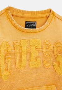 Sudadera amarilla de cuello redondo con logo bordado "GUESS" en un estilo texturizado y esponjoso. El cuello acanalado añade detalle al diseño de la tela.