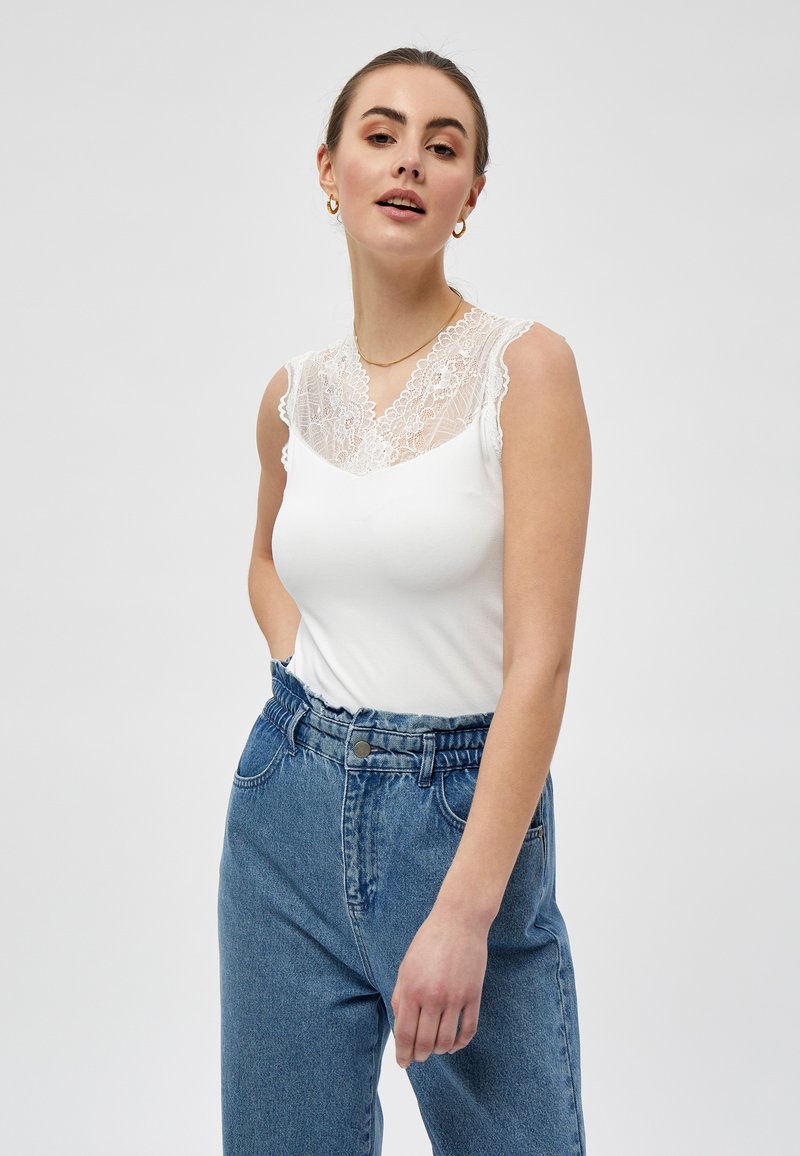 Top bianco senza maniche con dettagli in pizzo sul colletto, abbinato a jeans in denim blu a vita alta; presenta una vestibilità comoda e accenti in pizzo testurizzato.