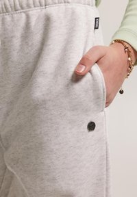 Harmaat fleece-housut ovat varustettu joustavalla vyötäröllä ja sivutaskulla, jossa on pieni musta logo. Kangas on pehmeä ja liukastuva.