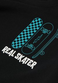 Černá látka s grafickým designem tří skateboardů v tyrkysové barvě, s kostkovaným vzorem, bleskem a textem "SKUTEČNÝ SKATER" pod nimi.