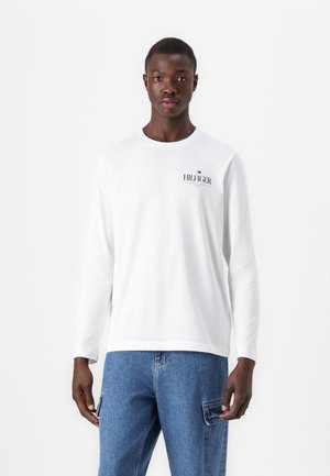 Long-sleeve white T-shirt made of cotton, featuring a small graphic logo in navy and red on the chest, worn with blue denim jeans.