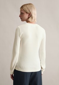 Langärmeliges, geripptes Oberteil in hellem Cremefarben, mit Rundhalsausschnitt, schlanker Passform und dezentem Seitentag-Detail. Weiche Textur mit glattem Finish.