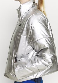 Person trägt eine glänzende silberne Steppjacke mit hohem Kragen, eine Hand in der Tasche, und darunter sind blaue, gemusterte Leggings sichtbar.