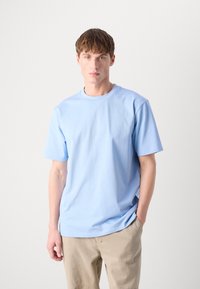 Ανοιχτό μπλε κοντομάνικο μπλουζάκι με στρογγυλή λαιμόκοψη, φτιαγμένο από μαλακό ύφασμα. Η εφαρμογή είναι χαλαρή, με εμφανή ραψίματα και χωρίς σχέδια.