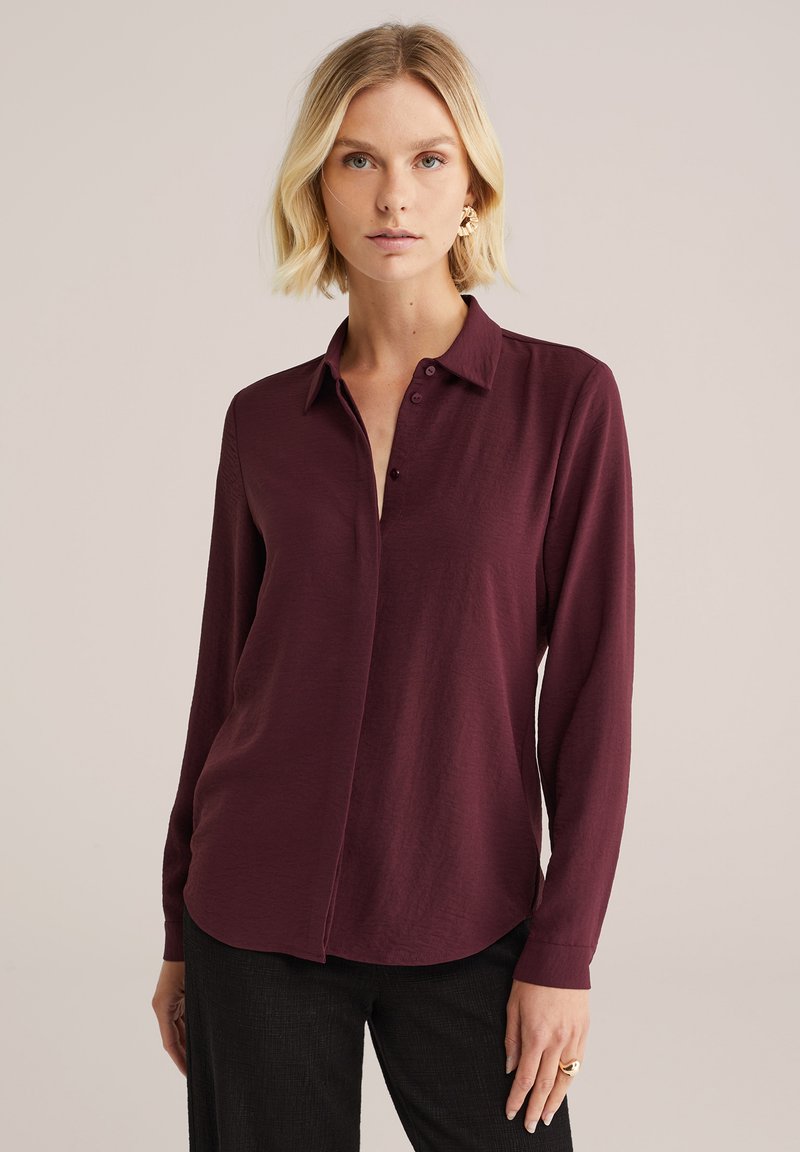 Camicia bordeaux a maniche lunghe con colletto, chiusura a bottoni, tessuto morbido e orlo arrotondato, abbinata a pantaloni scuri.