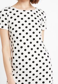 Šaty s krátkým rukávem z bílé látky s velkými černými puntíky, kulatým výstřihem a přiléhavým střihem.